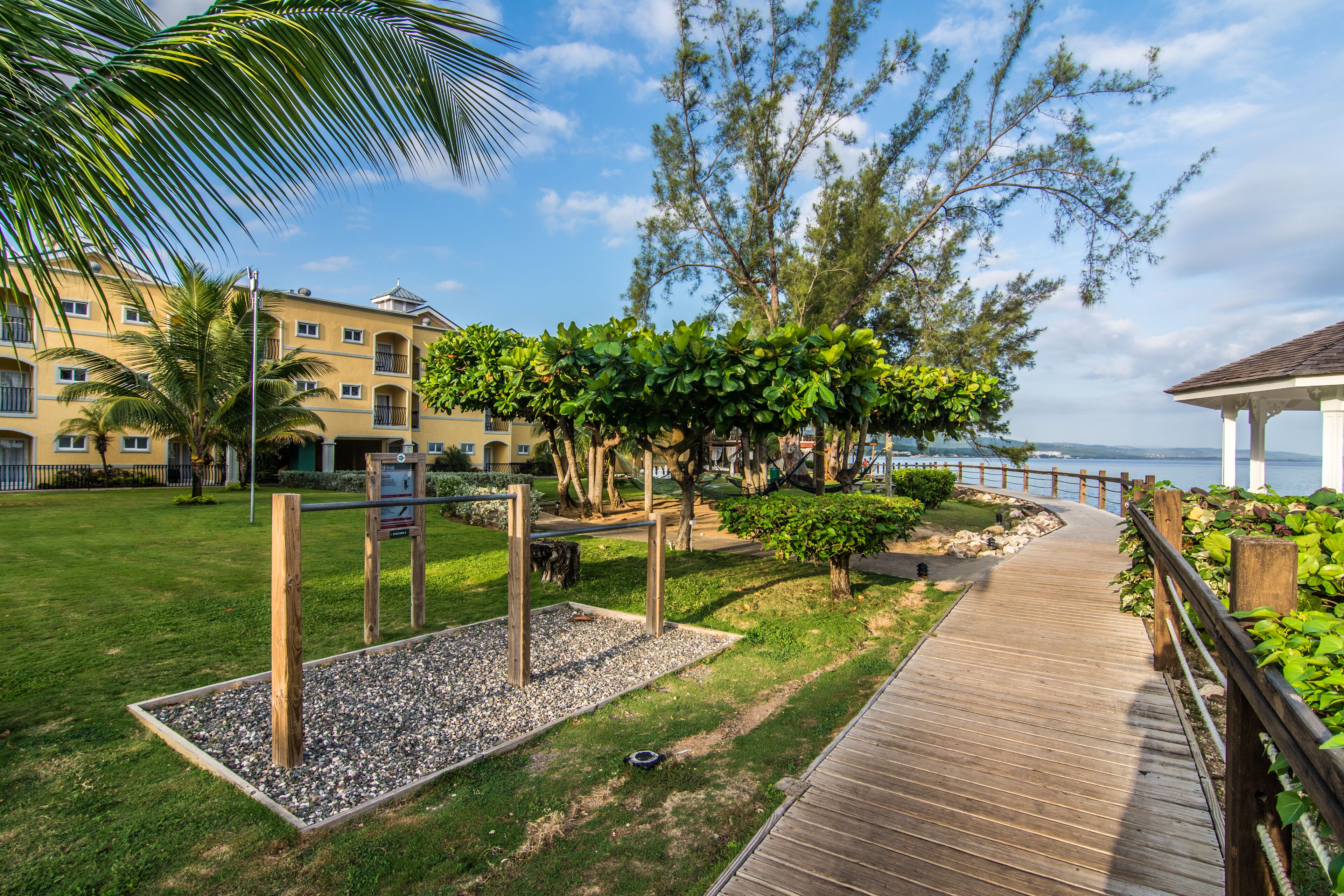 A wooden boardwalk leading to a gazebo with a view of the sea, surrounded by greenery and a building with balconies.