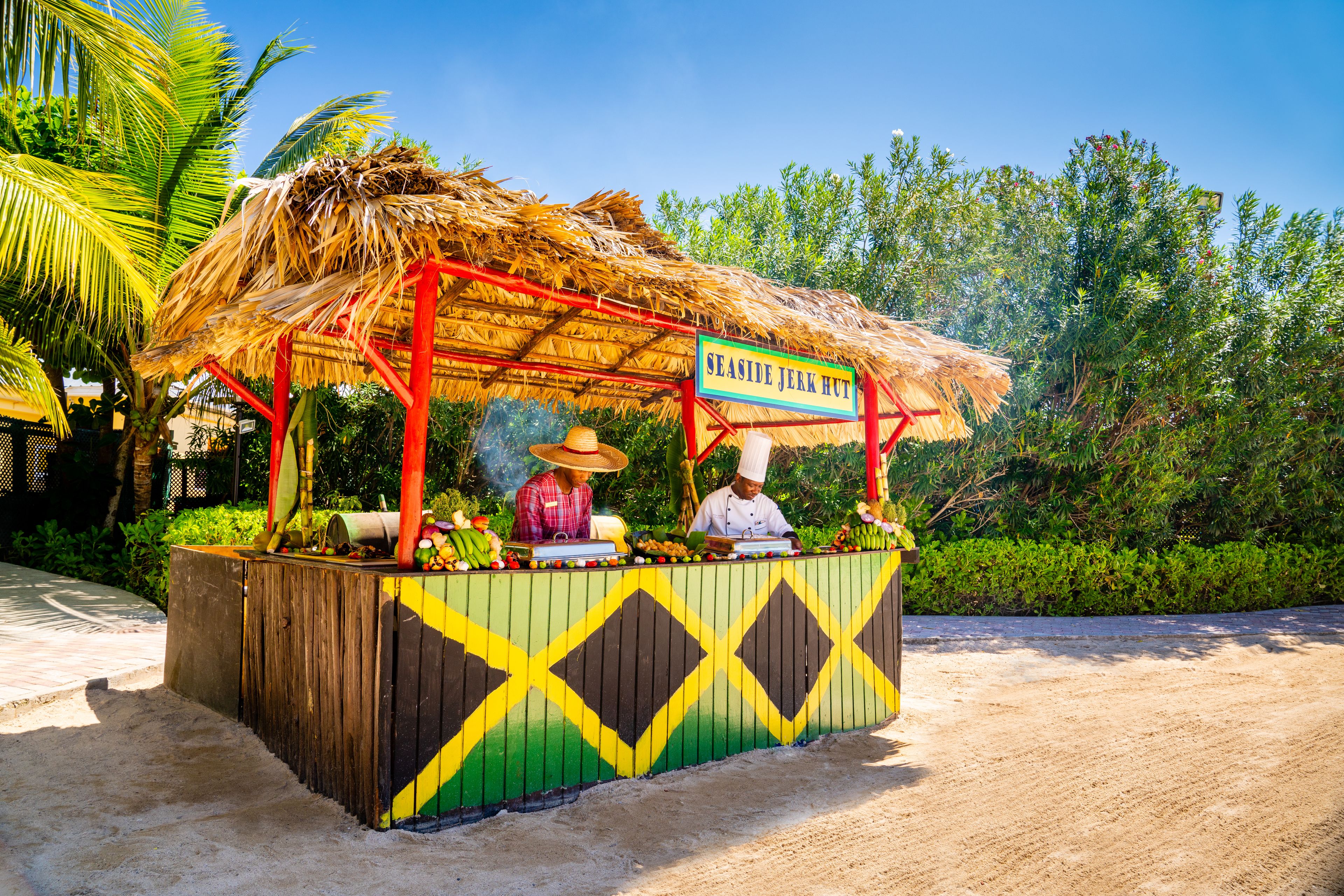 Un stand de restauration situé en bord de mer, appelé « Seaside Jerk Hut », avec un toit de chaume et une structure en bois aux couleurs vives.