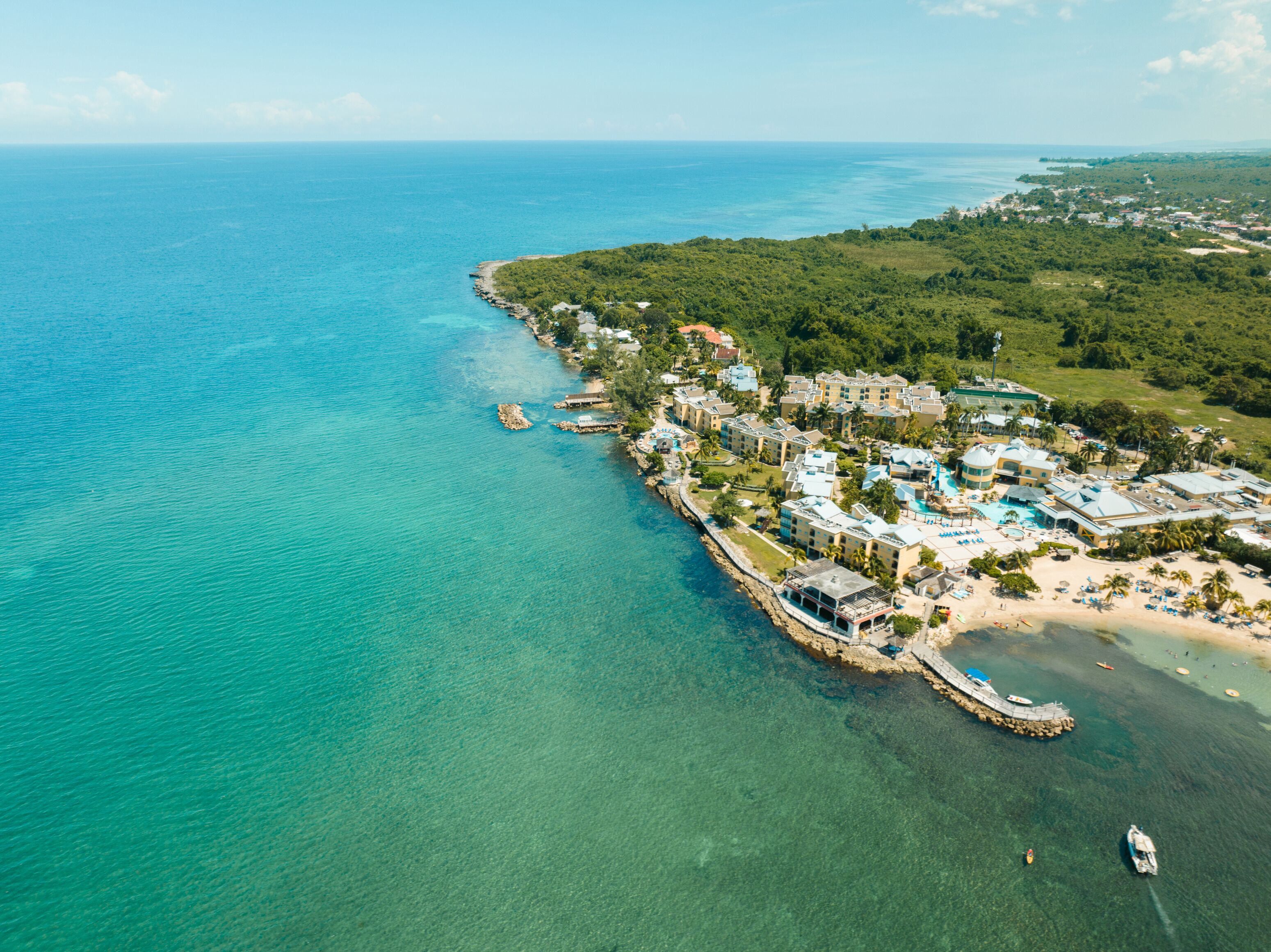 Vue aérienne d’une station balnéaire comprenant plusieurs bâtiments, une plage et des eaux d’un bleu limpide.