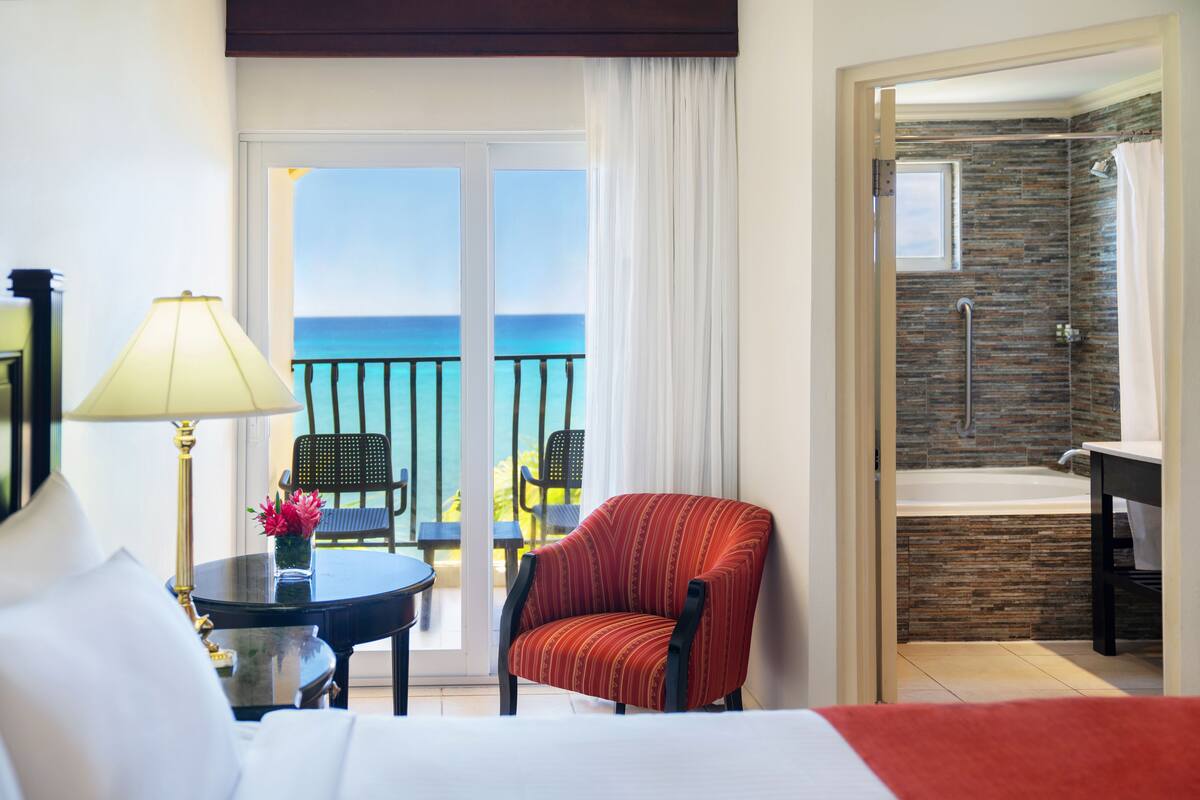 Une chambre d’hôtel comprenant un lit, une table avec une lampe, une chaise, un balcon avec vue sur la mer, et une salle de bain avec baignoire et douche.