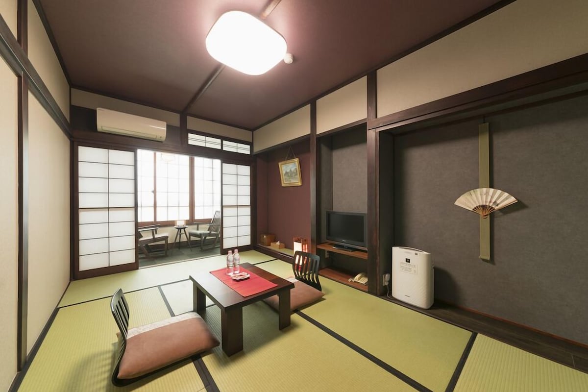Una habitación japonesa tradicional con suelo de tatami, una mesa baja, un televisor y un ventilador.