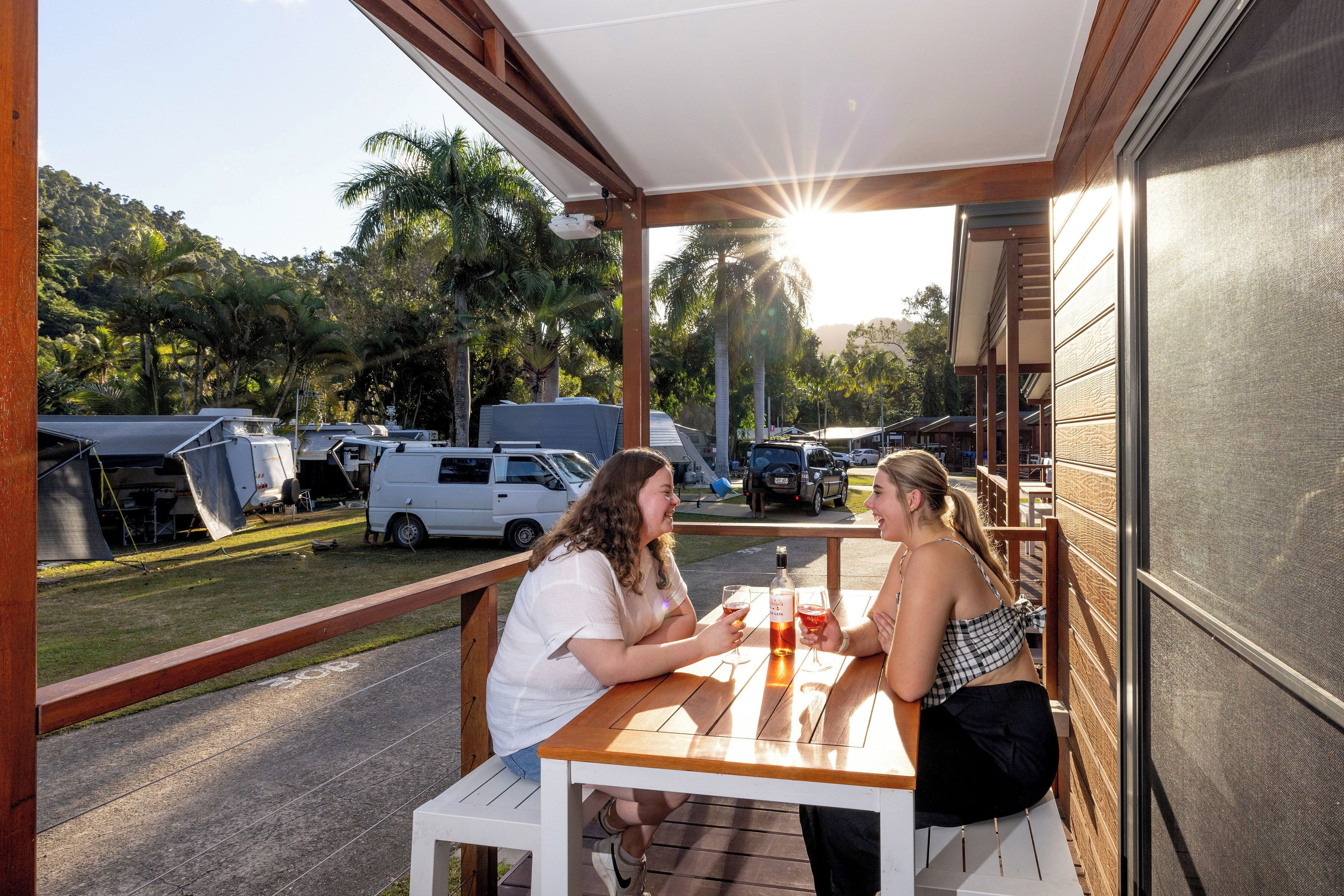 Un patio al aire libre con una mesa y bancos de madera, rodeado de una zona cubierta con vista a vehículos estacionados y palmeras al fondo.
