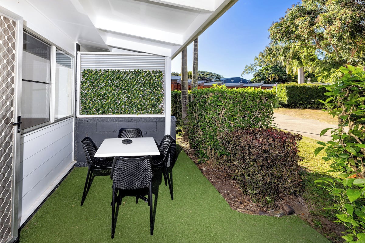 Une terrasse avec une table et des chaises, entourée d’un mur blanc et d’une haie.