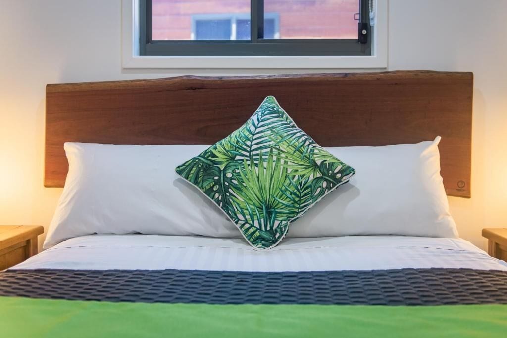 Una cama con cabecera de madera, almohadas blancas y un cojín con estampado de hojas verdes.