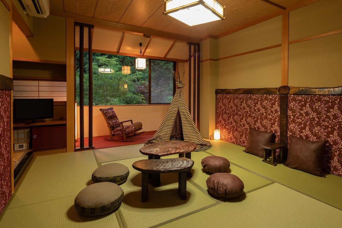 Um quarto japonês tradicional com tatames, uma televisão, uma cadeira de balanço, uma mesa pequena e vista para o verde lá fora.