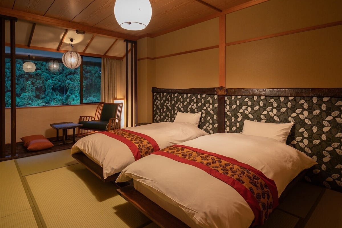 Um quarto em estilo tradicional japonês com piso de tatame, cama com cabeceira estampada, vista para uma floresta por uma porta deslizante e um pendente.