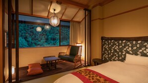 Habitación de hotel con una cama grande, cabecera de madera, ventana con vista a árboles, una silla y una mesita.