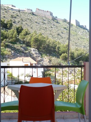 A balcony with a table and chairs, overlooking a mountainous landscape with a historical fortification. - Hotel Vasilis (Nafplio)