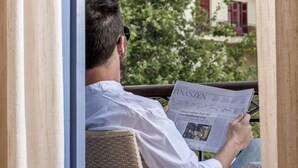 A man reading a newspaper in a wicker chair with a view of a building and trees. - Xenon Inn (Nafplio)