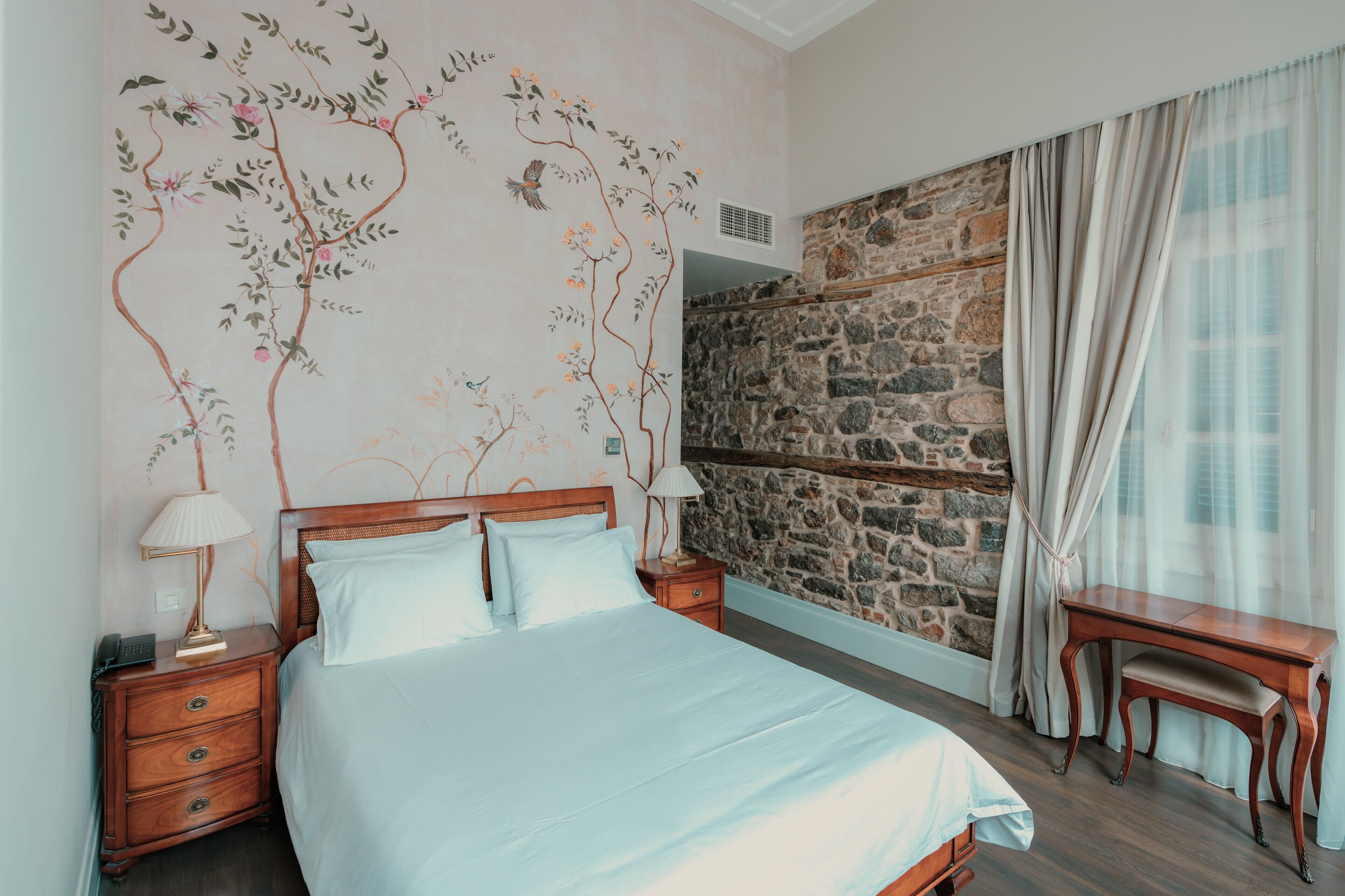 Une chambre à coucher avec un lit, des tables de chevet, un bureau, un mur en pierre et une fenêtre avec des rideaux.
