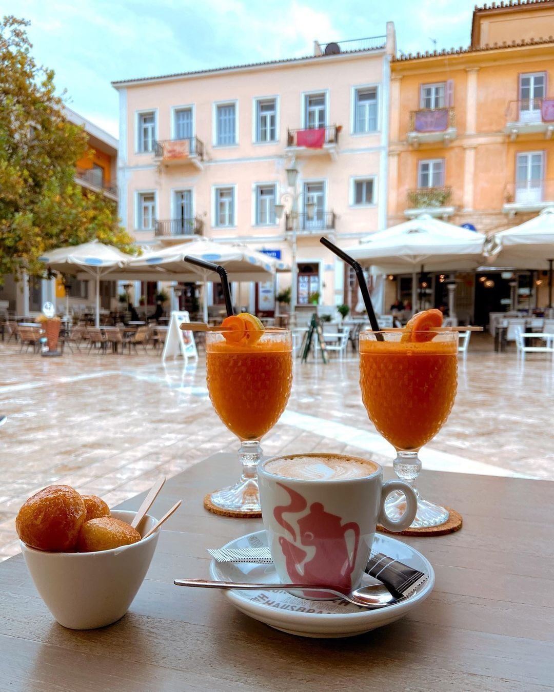 Uma mesa com dois copos de bebida de laranja, uma chávena de café e uma travessa de pastelaria, disposta ao ar livre com um edifício ao fundo.