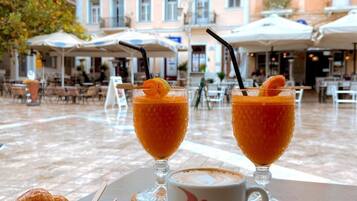 A table with two glasses of orange beverage, a cup of coffee, and a bowl of pastries, set outdoors with a building in the background.