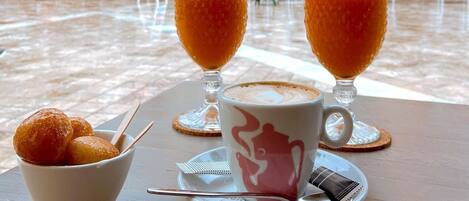Uma mesa com dois copos de bebida de laranja, uma chávena de café e uma travessa de pastelaria, disposta ao ar livre com um edifício ao fundo.