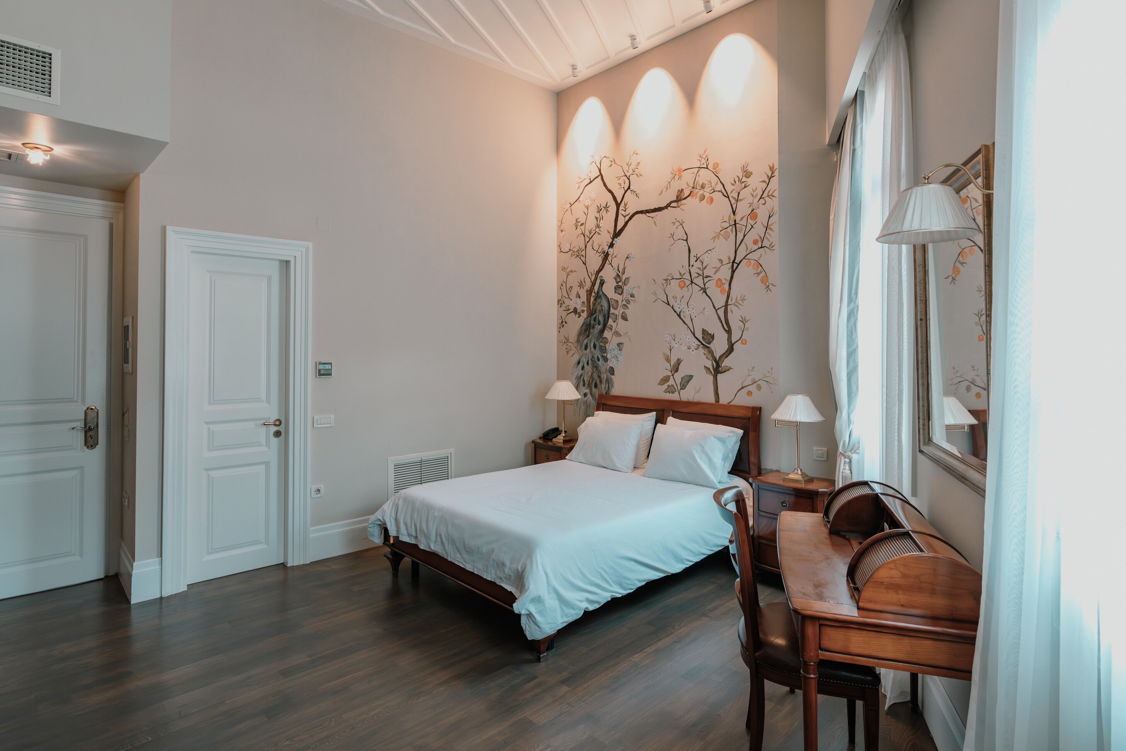 Une chambre à coucher comprenant un lit, des tables de chevet, un bureau et une chaise. Il y a deux portes et une fenêtre avec des rideaux.