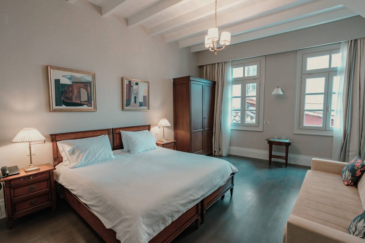 A hotel room with a large bed, bedside tables with lamps, a wardrobe, a small table, and a sofa.