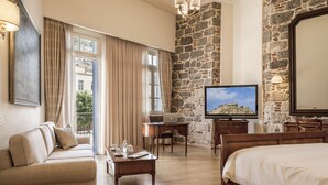 A hotel room with a stone wall, a bed, a sofa, a desk with a laptop, a television, and a balcony with curtains. - Xenon Inn (Nafplio)