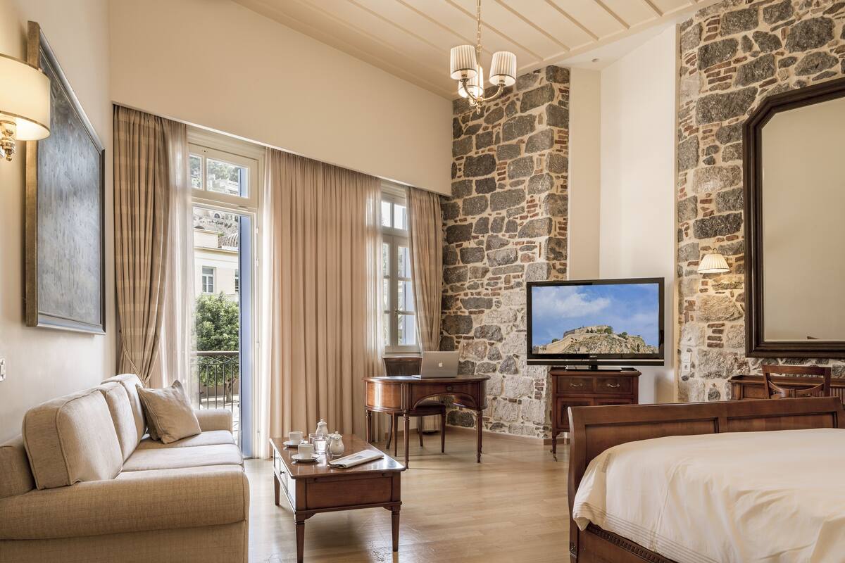 A hotel room with a stone wall, a bed, a sofa, a desk with a laptop, a television, and a balcony with curtains.