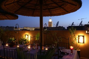 Un área de comedor al aire libre con mesas dispuestas para una comida, rodeada de plantas en macetas y cubierta por un toldo de paja.