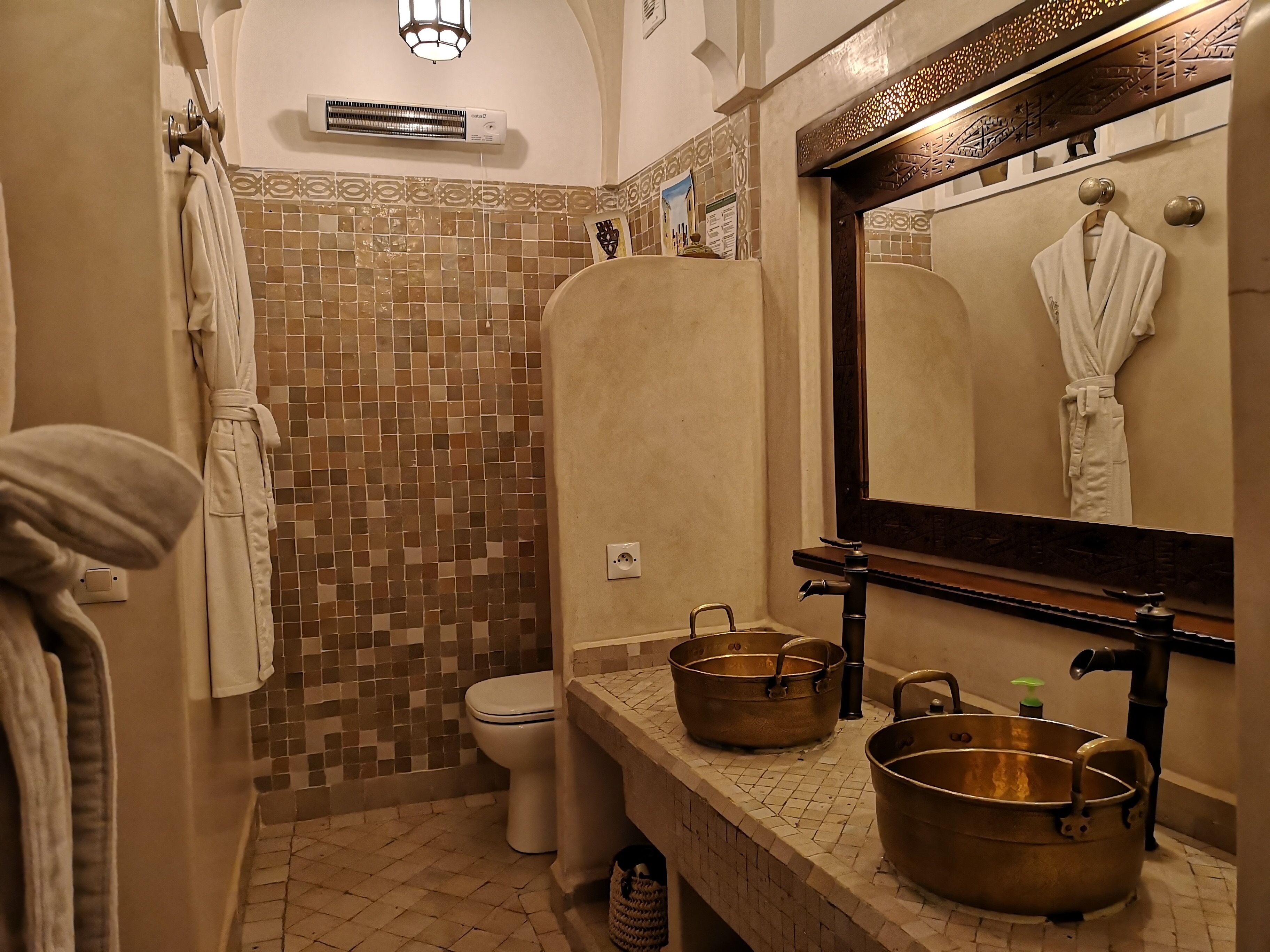 A bathroom with a tiled shower wall, a toilet, a sink with two copper pots, and a mirror with a robe hanging on it.