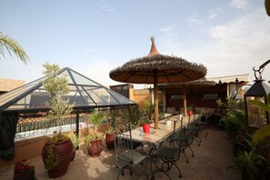 Un patio al aire libre con una mesa larga y sillas, rodeado de plantas en macetas y un paraguas grande.