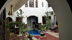 Ein Innenhof mit Pool, umgeben von Kakteen und Topfpflanzen, mit Blick auf ein weißes Gebäude mit Bogenfenstern und Fliesenboden.