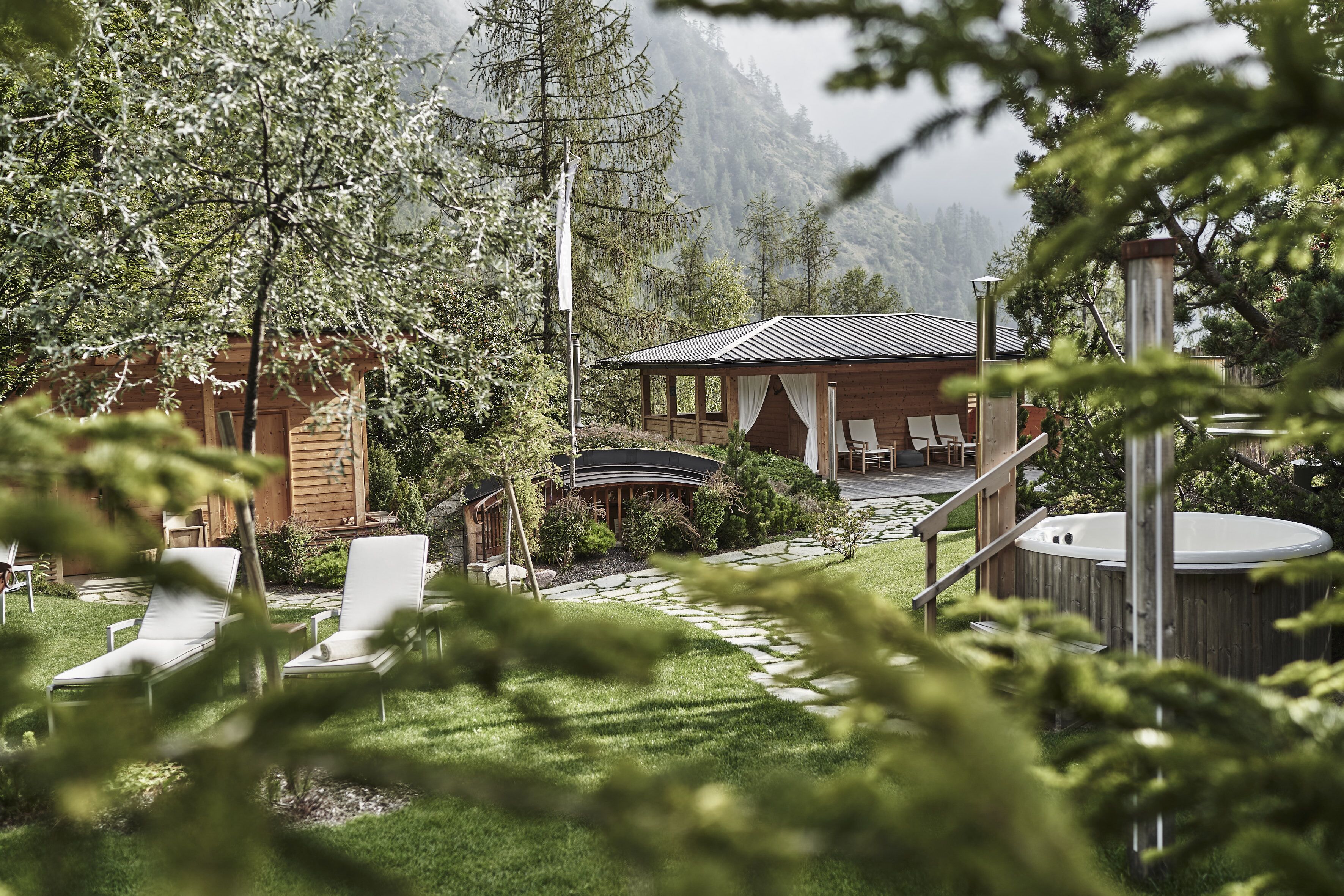Eine Holzhütte mit Whirlpool, Liegestühlen und einer überdachten Terrasse, umgeben von Bäumen und Bergen.