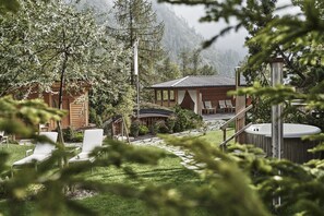A wooden cabin with a hot tub, lounge chairs, and a covered patio area surrounded by trees and mountains.
