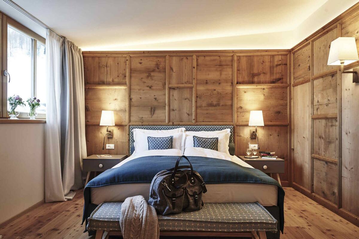 Une chambre avec une tête de lit en bois lambrissé, un lit avec une literie bleue, deux lampes de chevet, un banc avec un sac, et une fenêtre avec des rideaux.