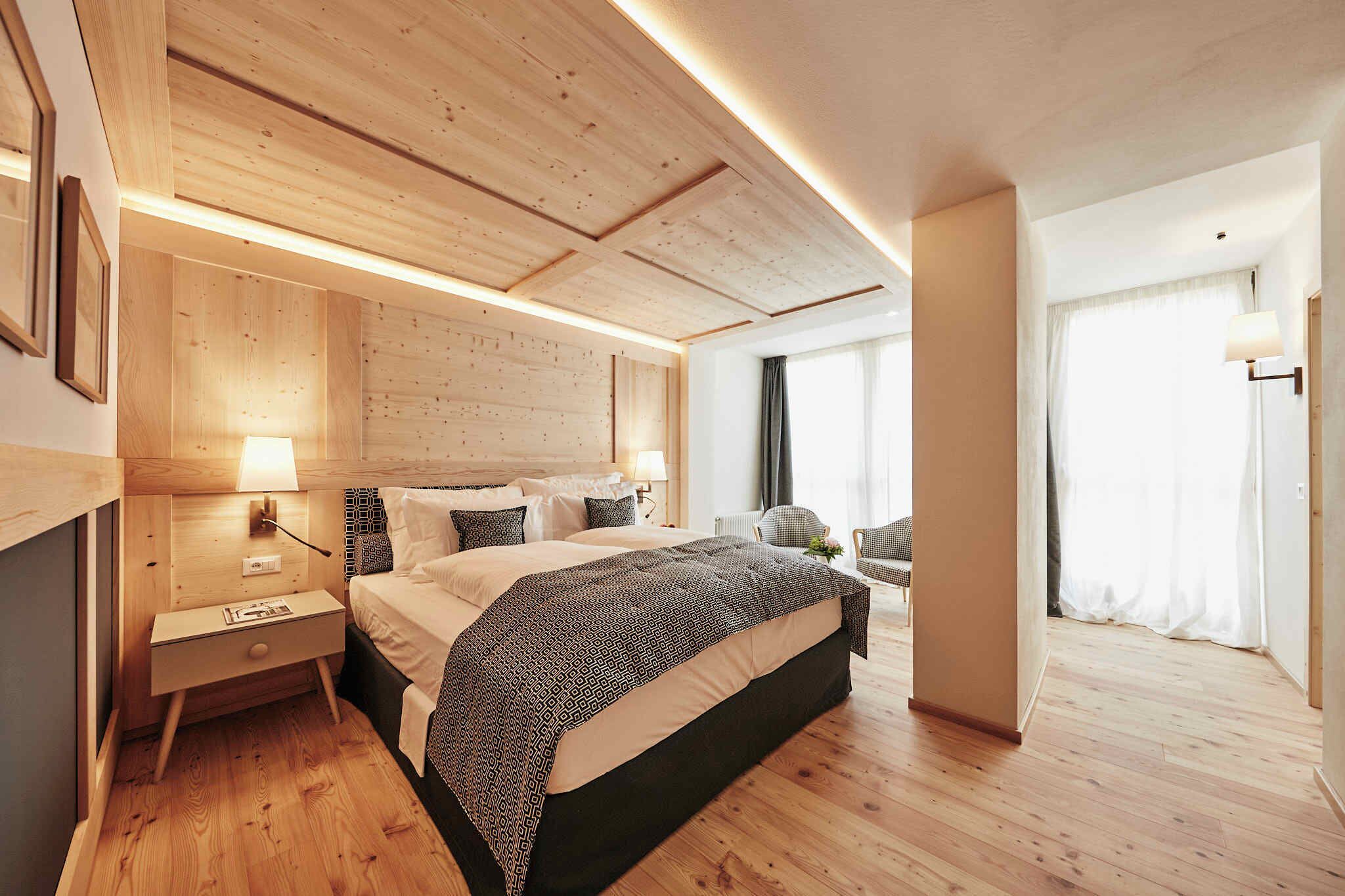 Ein modernes Hotelzimmer mit Holzboden, einem großen Bett mit gemusterten Textilien, einem Nachttisch mit Lampe und Blick auf einen Balkon mit Vorhängen.