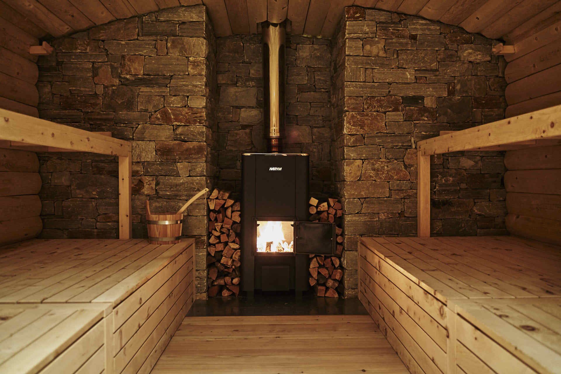 A log cabin with a stone wall interior, a central wood-burning stove, and wooden benches.