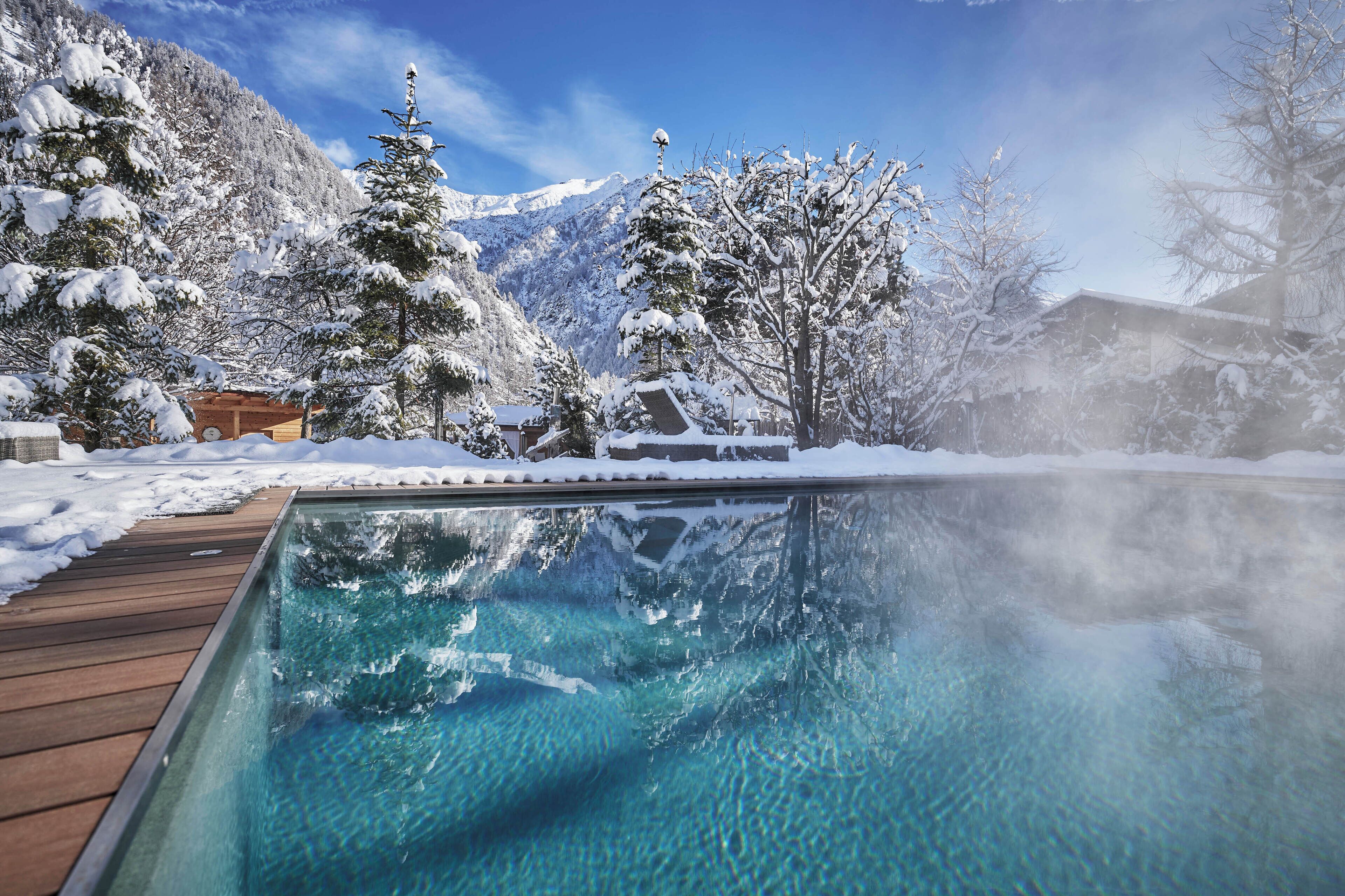 Una piscina cristalina con cubierta de madera, rodeada de árboles nevados y montañas al fondo.