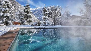 Una piscina cristalina con cubierta de madera, rodeada de árboles nevados y montañas al fondo.