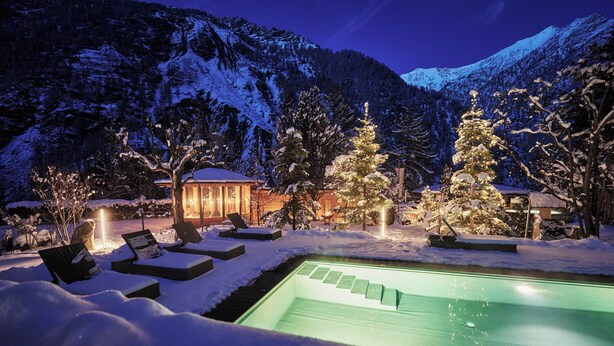 A snow-covered outdoor area with a pool, lounge chairs, and illuminated trees, set against a mountainous backdrop. - Goldene Rose Karthaus (Senales)