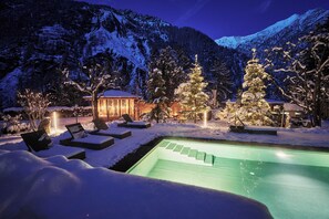 Ein verschneiter Außenbereich mit Pool, Liegestühlen und beleuchteten Bäumen, vor einer Bergkulisse.