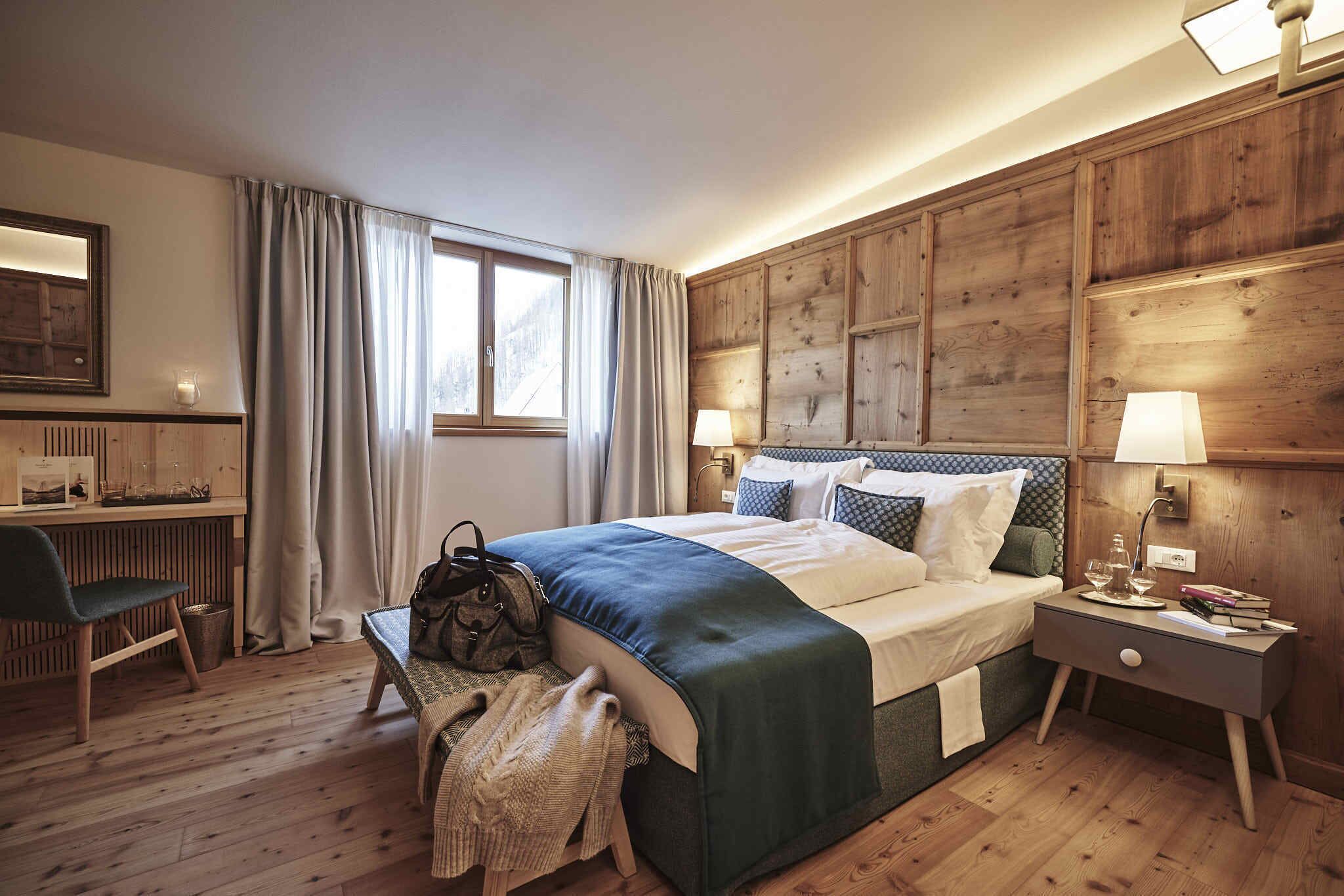 A hotel room with a wooden paneled wall, a bed with a blue and white bedspread, a bedside table with a lamp, a chair, and a desk with a lamp.
