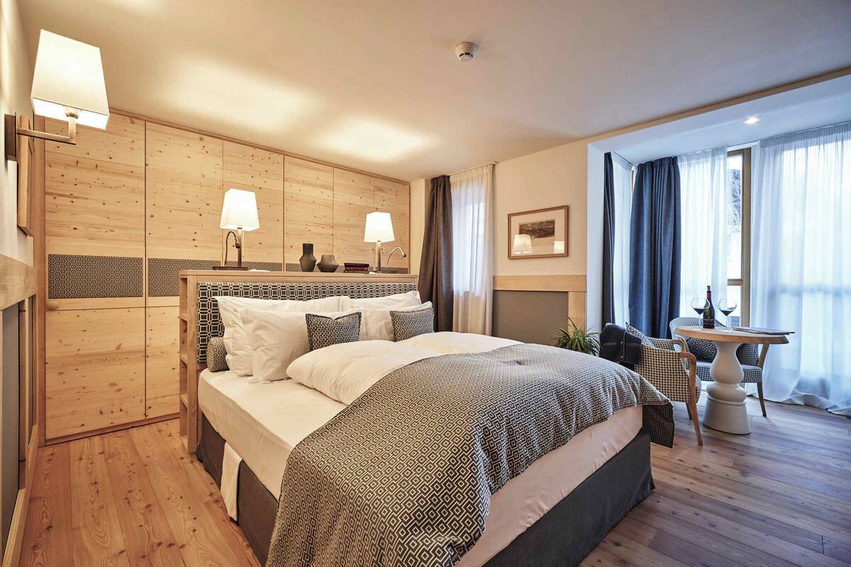 Une chambre d’hôtel moderne avec un grand lit, des lambris en bois sur les murs, une petite table avec une chaise et une fenêtre avec des rideaux.