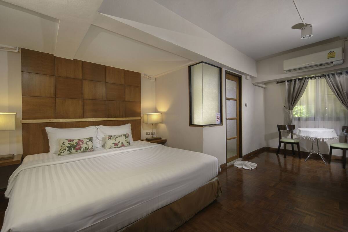 A hotel room with a large bed, a wooden headboard, a bedside table with a lamp, a small table with chairs, and a window with curtains.