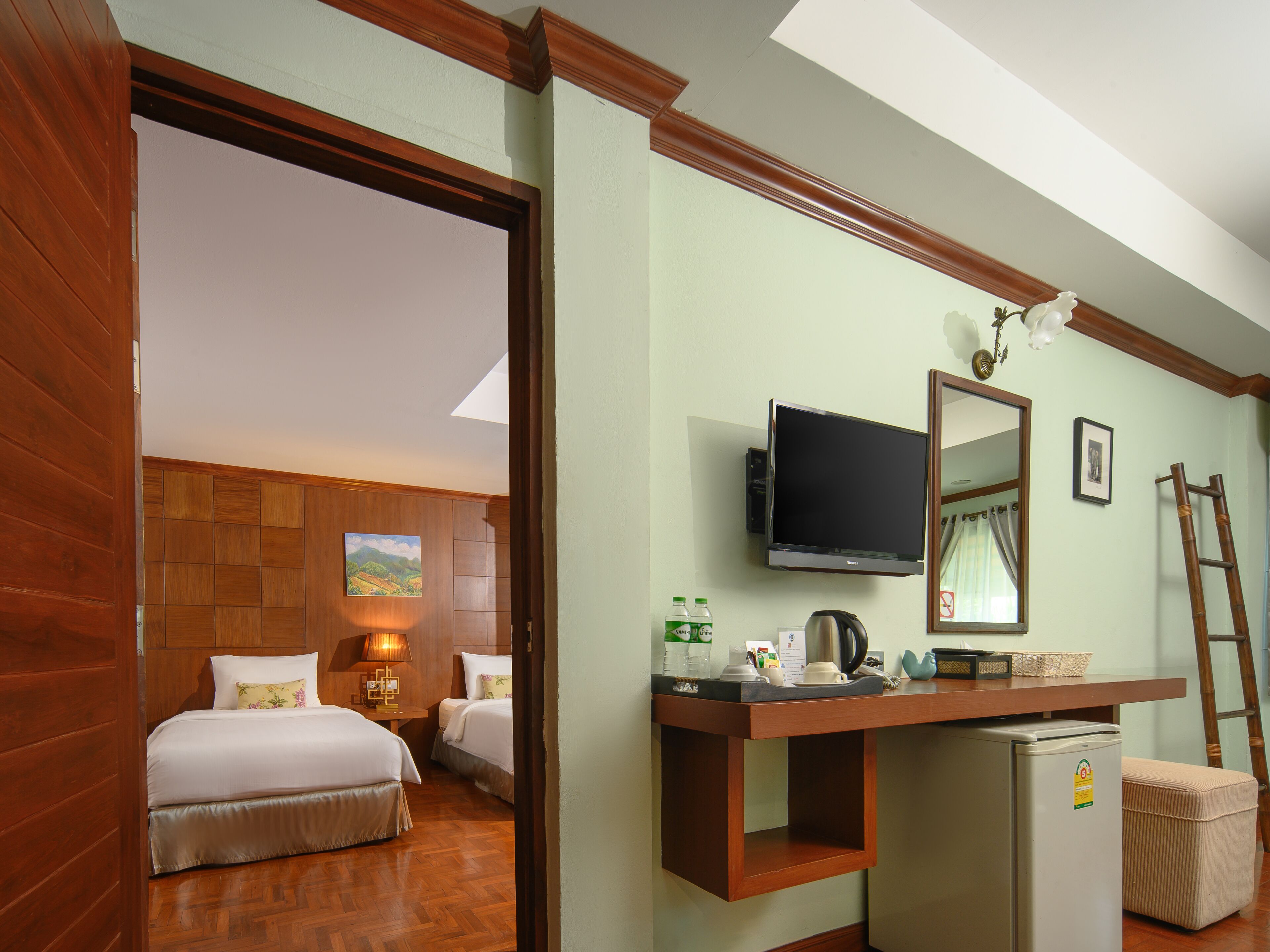 A hotel room with two beds, a television, a refrigerator, a desk with a mirror, and a wooden floor.