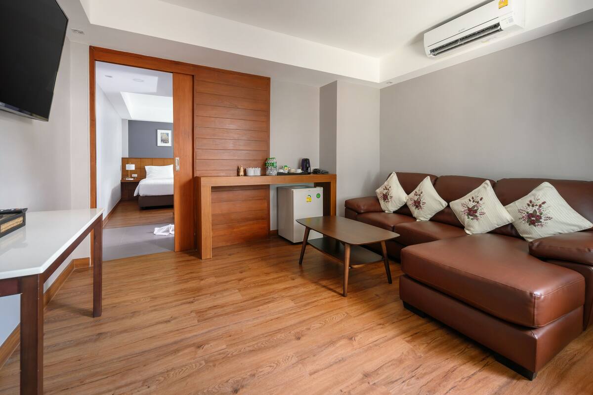 A modern hotel room with a wooden floor, a brown leather sofa with floral cushions, a small wooden table, a TV, and a kitchenette area.