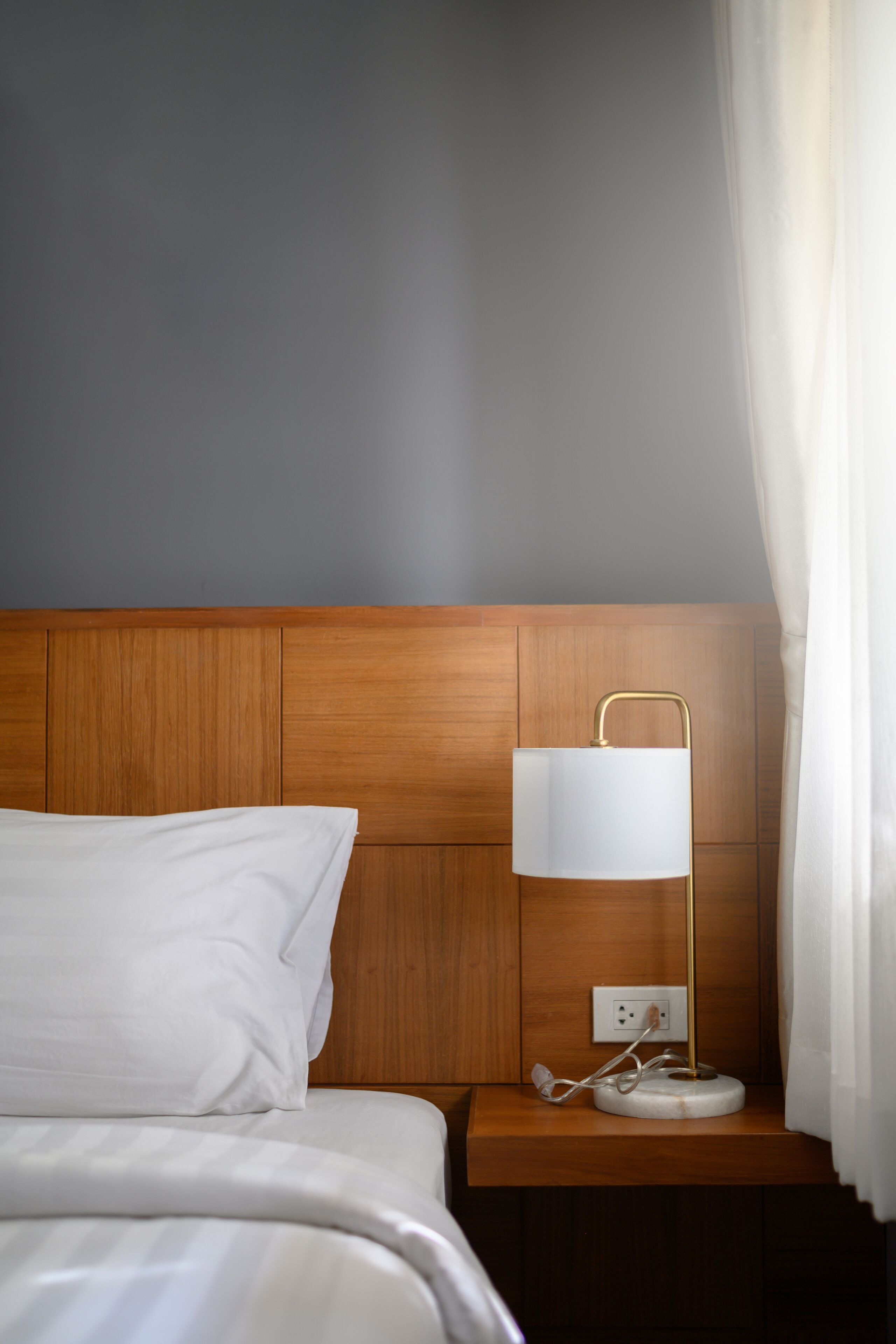 A neatly made bed with white linens, a wooden headboard, a bedside table with a lamp, and a power outlet.
