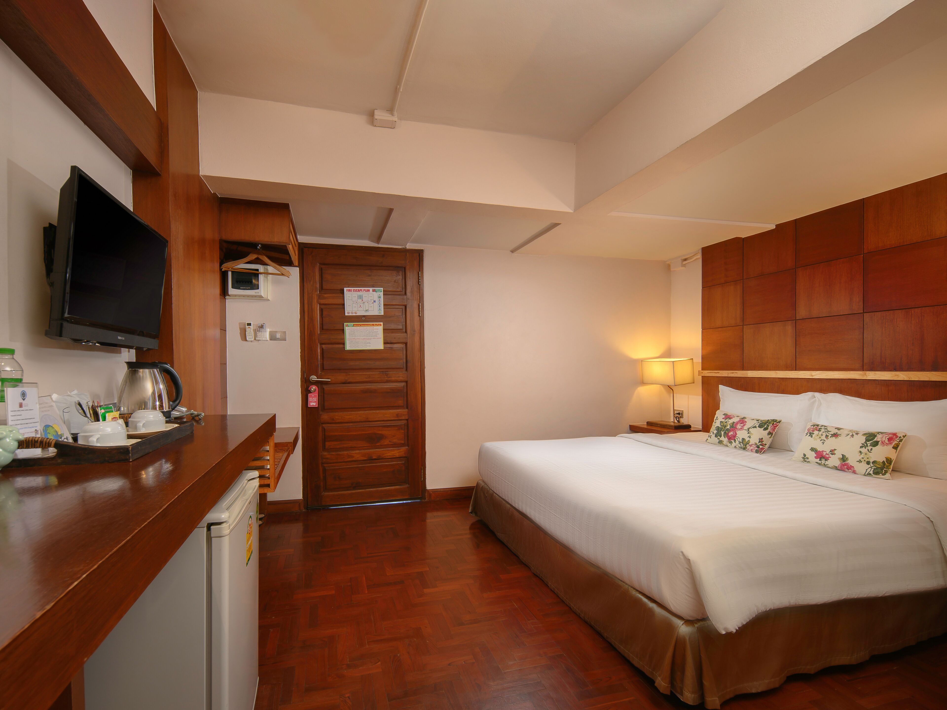 A hotel room with a wooden headboard, a bed with white bedding, a bedside lamp, a wooden desk with a television, and a coffee maker.