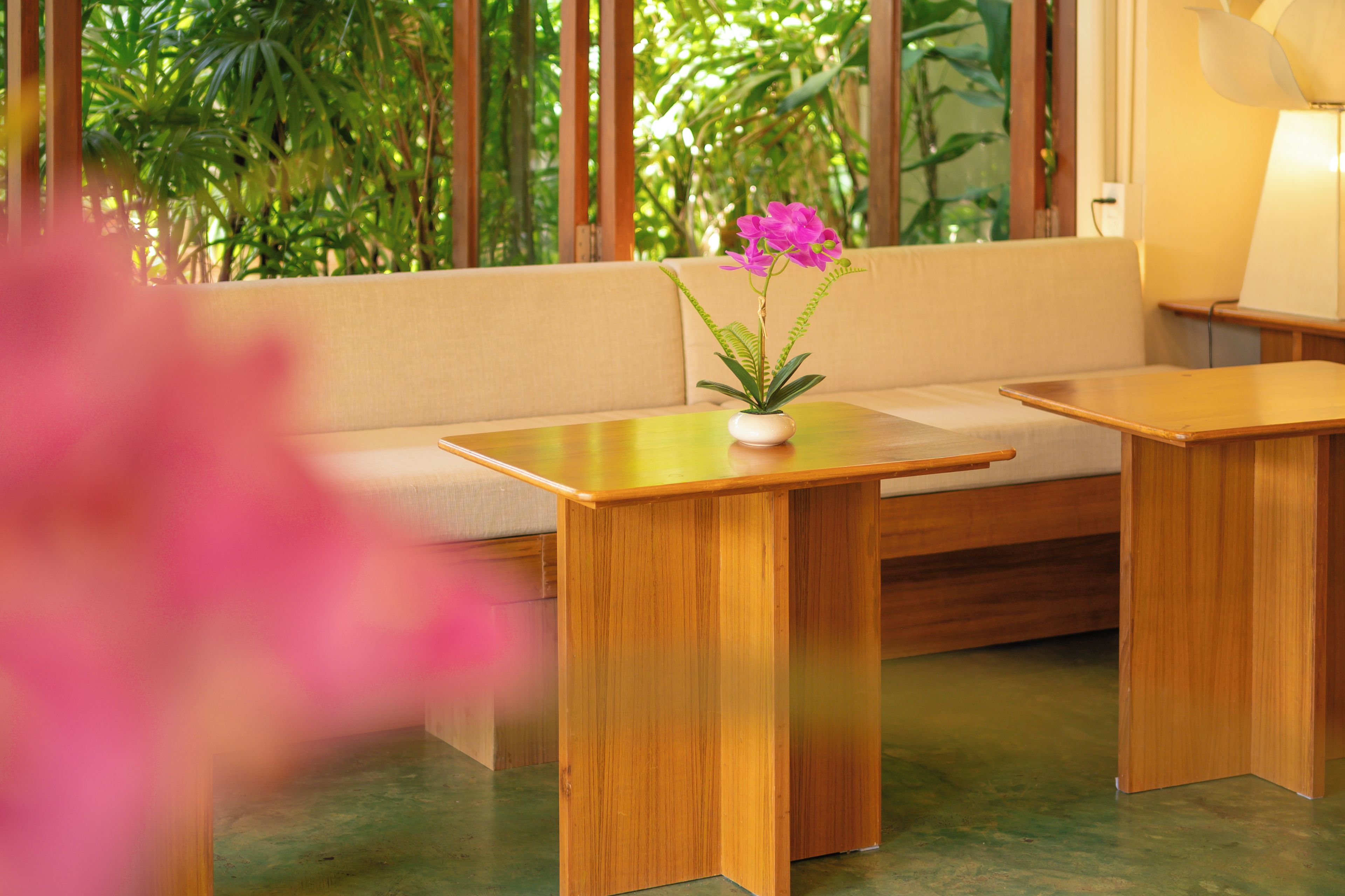 A room with a wooden bench, a small table with a vase of flowers, and a view of greenery through large windows.