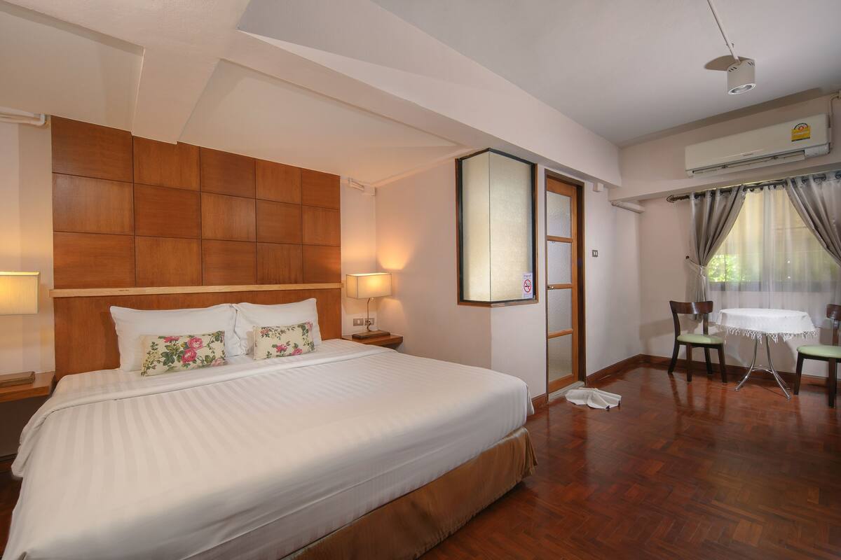 A hotel room with a large bed, a wooden headboard, a small table with chairs, and an air conditioning unit.