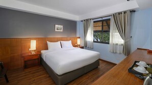 Habitación de hotel con una cama grande, cabecera de madera, dos mesitas de noche con lámparas, un escritorio de madera con una bandeja de tazas de té y una ventana con cortinas.