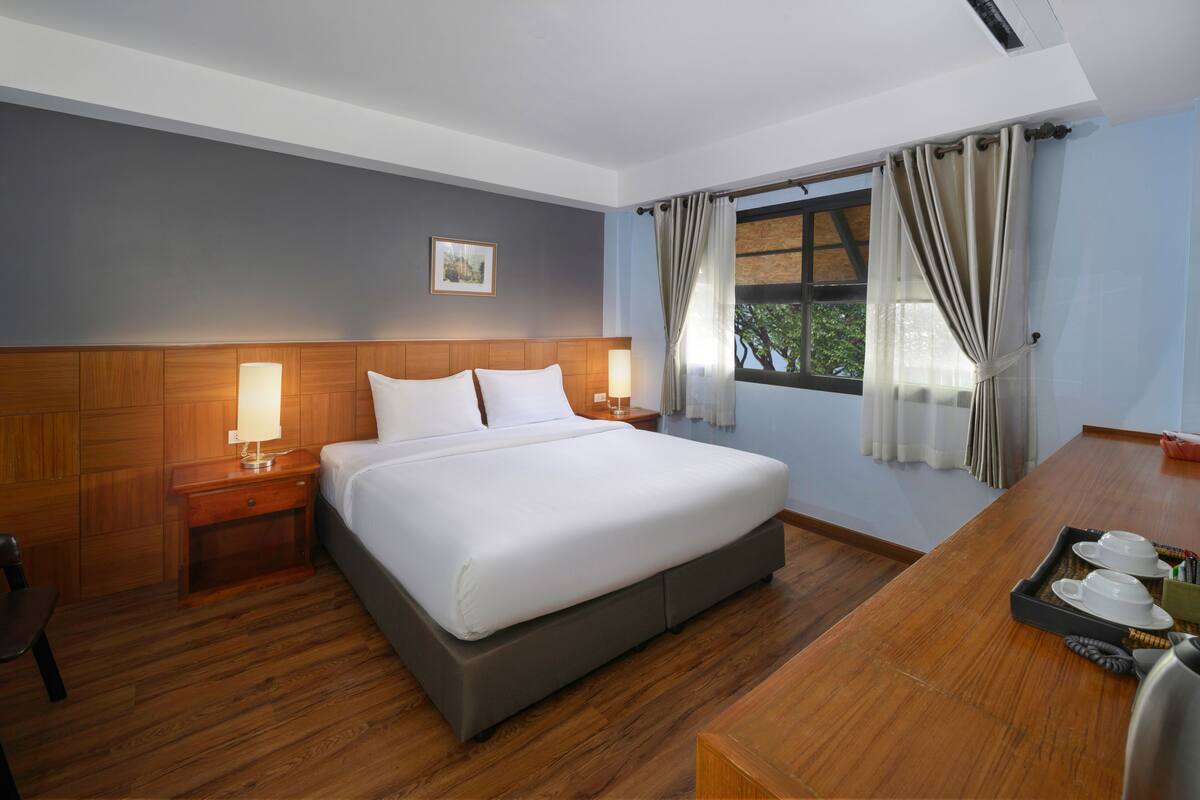 A hotel room with a large bed, wooden headboard, two bedside tables with lamps, a wooden desk with a tray of teacups, and a window with curtains.