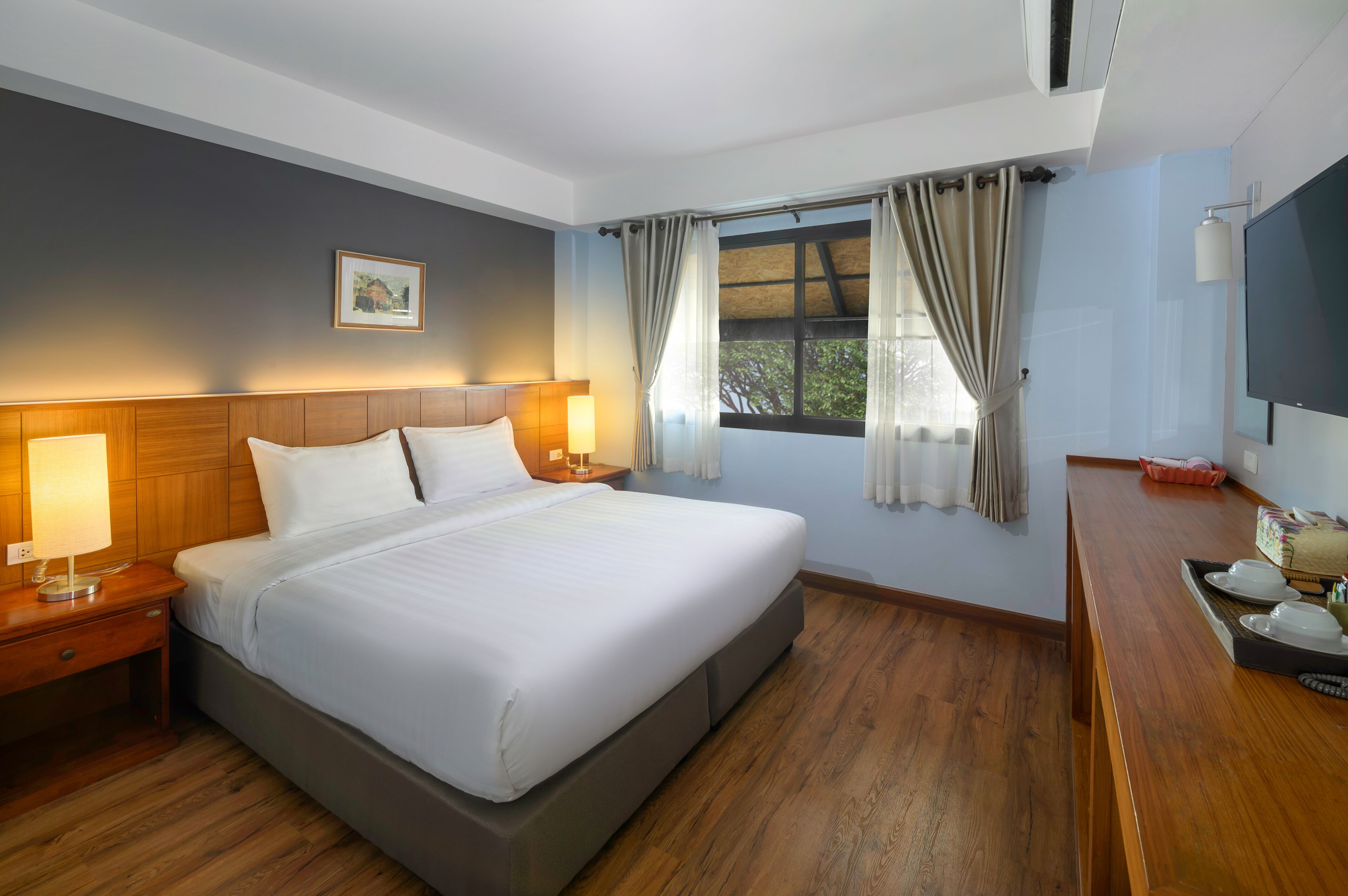 A hotel room with a large bed, wooden headboard, bedside lamps, a flat-screen TV, and a window with curtains.