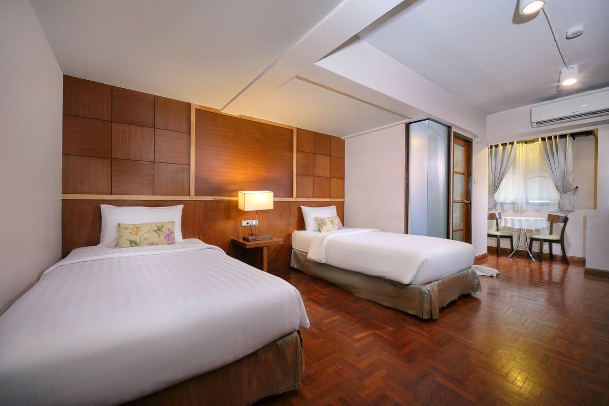 A hotel room with two beds, wooden headboards, a small table with chairs, and a window with curtains.