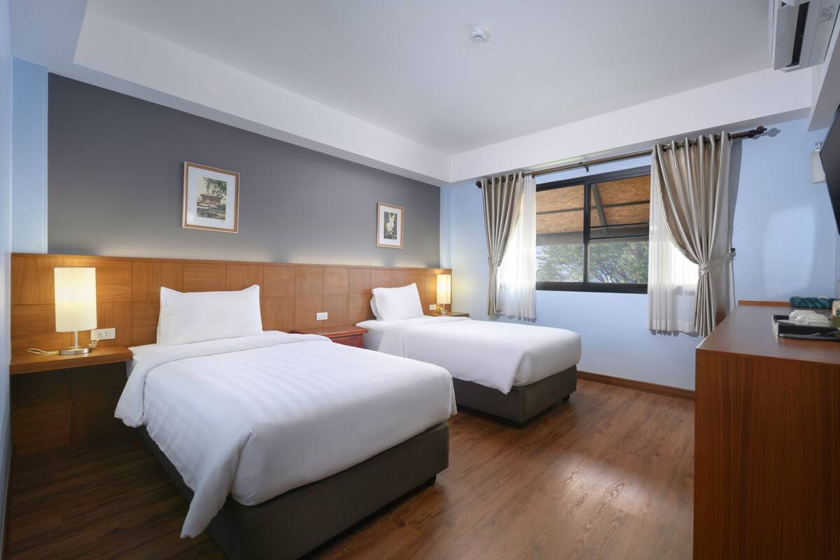 A hotel room with two beds, wooden headboard, bedside tables with lamps, a desk, and a window with curtains.
