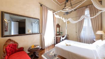 Habitación de hotel con una cama grande, un sillón rojo, una mesita de noche de madera, un espejo y una ventana con cortinas.