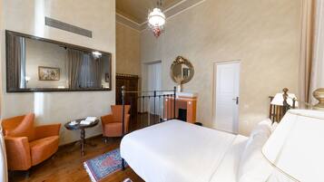 A hotel room with a large bed, two armchairs, a small table, a mirror, a dresser, and a chandelier.